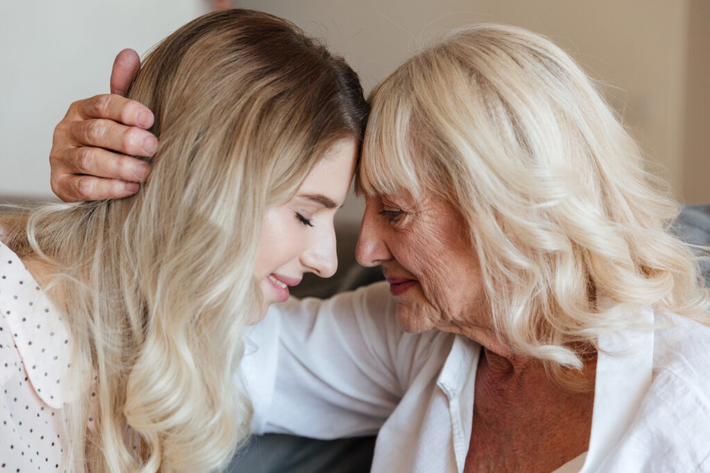 Picture of smiling young lady sitting at home with her grandmother hugging. Looking aside.