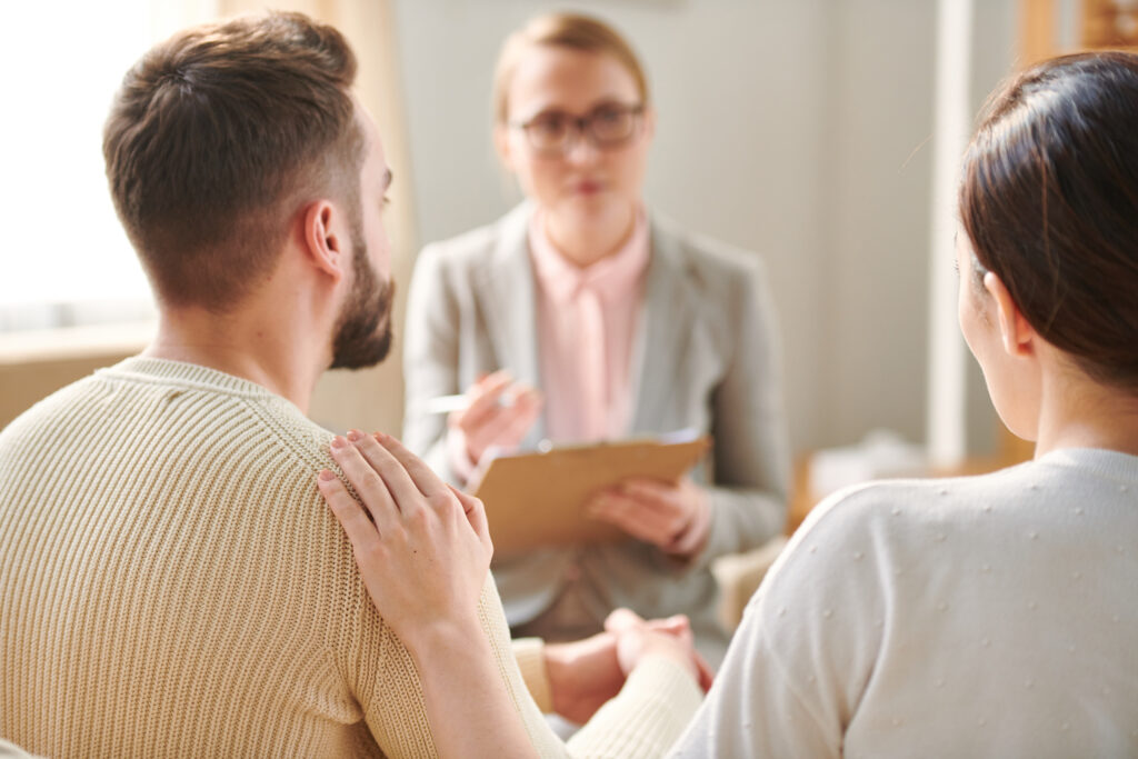 Rear view of young wife keeping hand on her husband shoulder while both sitting in front of psychotherapist during consultation